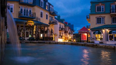 Scenic evening at Mont-Tremblant Resort pedestrian village showing illuminated luxury lodging and a water feature in the central square