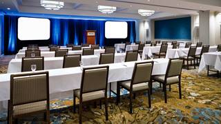 A large, professional conference room set up with long white tables, padded chairs, and dual projection screens against blue draped walls.