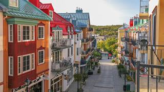 A high-angle view looking down a cobblestone street in the Mont-Tremblant pedestrian village, lined with colorful multi-story buildings and balconies.