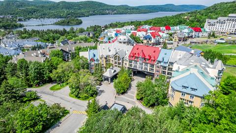 Aerial summer view of the Sommet des Neiges hotel at Tremblant Resort with colorful rooftops, green trees, and Lake Tremblant in the distance.