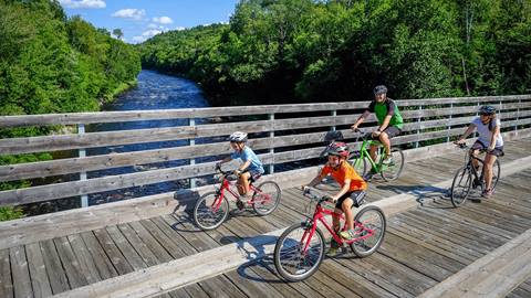 Famille à vélo traversant un pont de bois au-dessus d’une rivière, entourée de forêt, sur le sentier reliant le Domaine Saint-Bernard