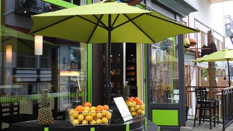 Exterior of Fluide Bar à Jus in the Mont-Tremblant pedestrian village featuring a green umbrella and fresh fruit displays of oranges and pineapples.